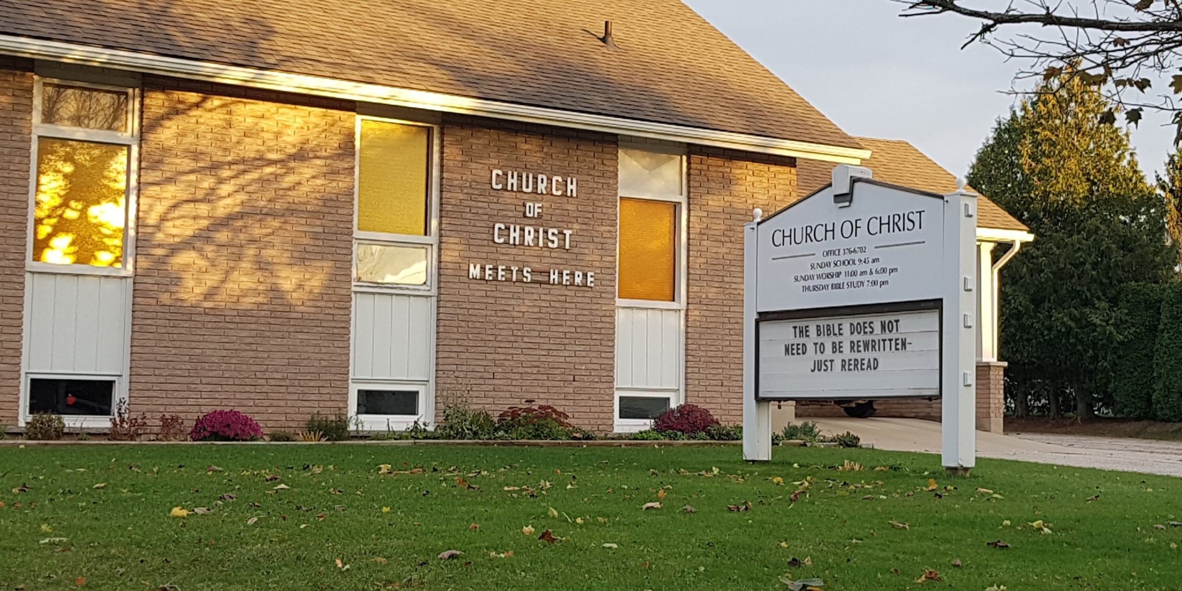 The low brick building where the Church of Christ meets, lit from within at dusk, with a sign on the lawn reading Church of Christ Meets Here