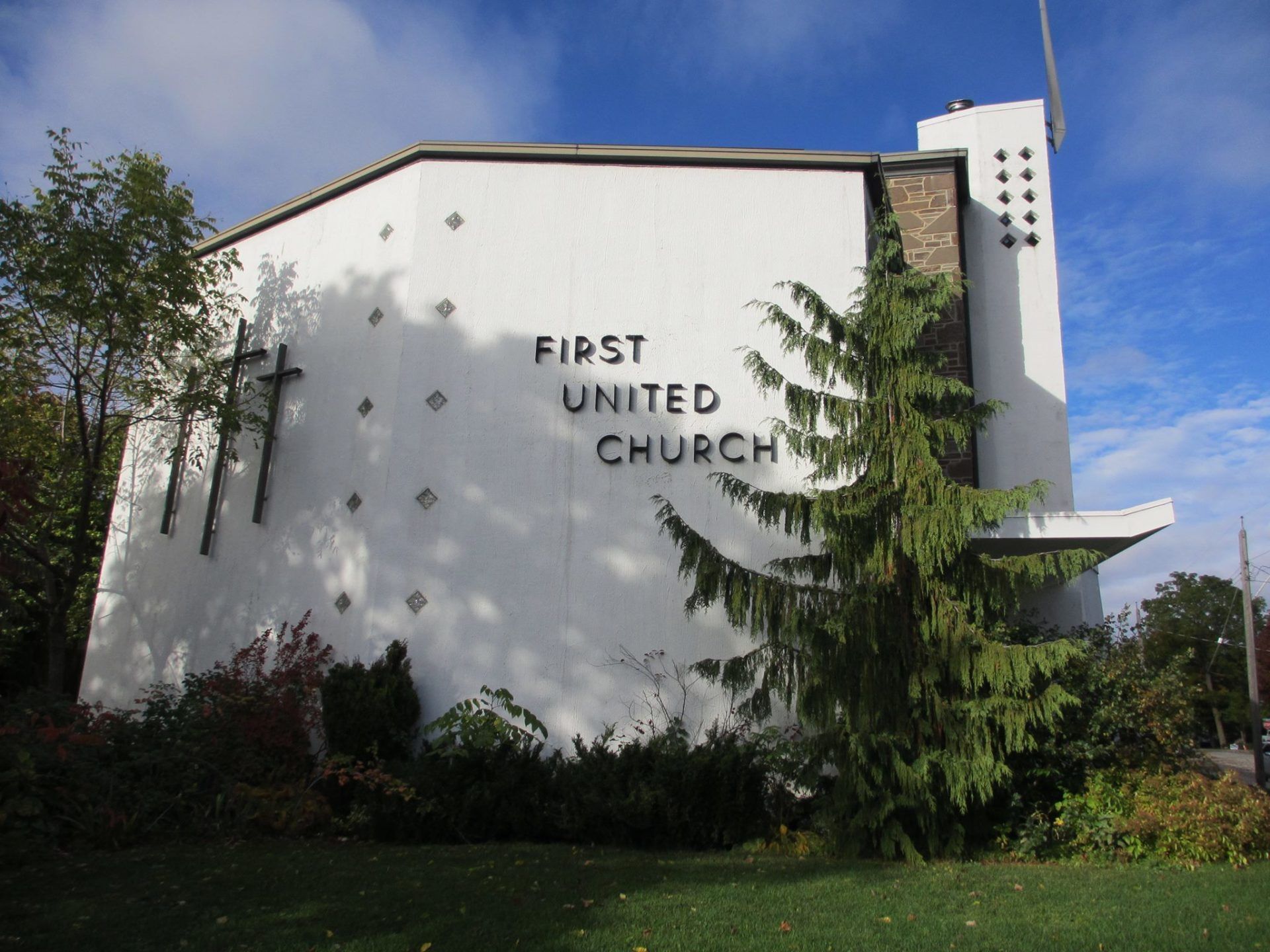 First United Church, a white modernist church with three crosses and its name lettered on the end wall, behind evergreens